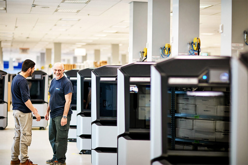 Två tekniker i en industriell produktionsmiljö med rad av 3D-skrivare för additiv tillverkning och prototyptillverkning.